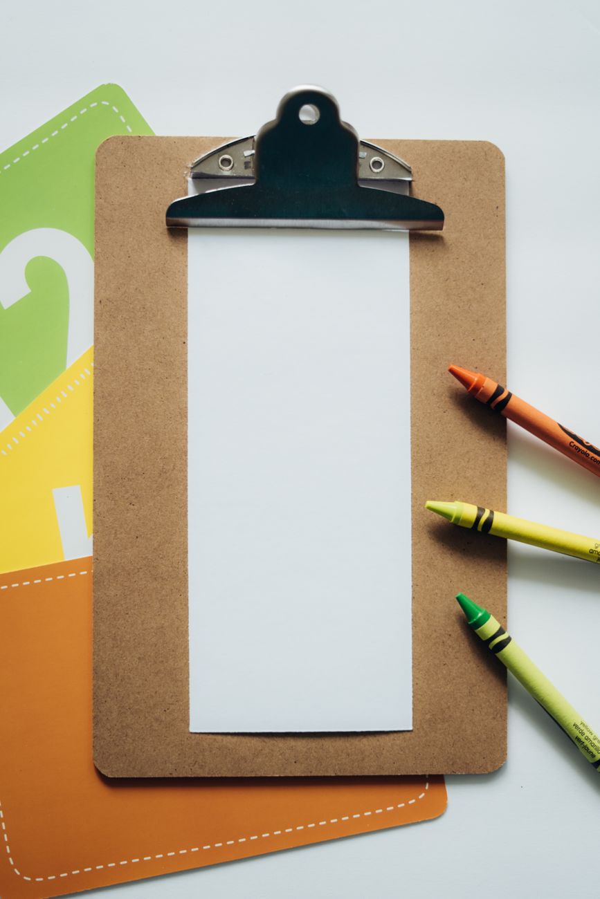 A clipboard with a blank sheet of paper, flanked by green, yellow, and orange crayons, on a white surface with colorful paper cards in the background.
